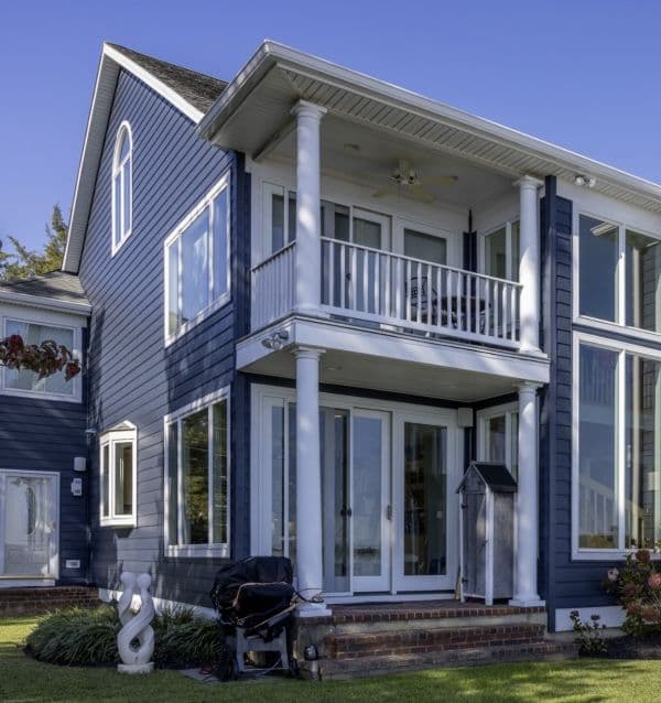 Blue house with large windows, white balcony, columns, and patio with grill under clear sky